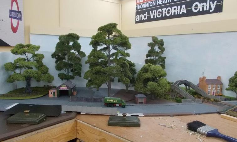 Trompe l'oeil ( Deceive the Eye').
At the London-end of Ewhurst Green, trains will pass under the footbridge before disappearing from view. This is achieved by a curved board in front of the track; itself obscured by trees. 
Six trees have been positioned on this curved board with work progressing (left to right) on the groundworks (foliage) which will soften up the skyline at ground level. A seventh tree is beyond the railway line; part of an eighth is just visible in a front garden.
In reality the sky is far ‘less’ blue that it appears here in these photographs.
www.EwhurstGreen.com 
