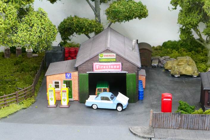 Ewhurst Green’s village garage is typical of this period; this being a Bachmann Scenecraft 44-076 model with much detail added around the garage’s yard. 
A 1959 Triumph Herald 948 Coupé receives attention while the rear of a 1960 Morris Mini Traveller can just be seen inside the garage.  
www.EwhurstGreen.com
