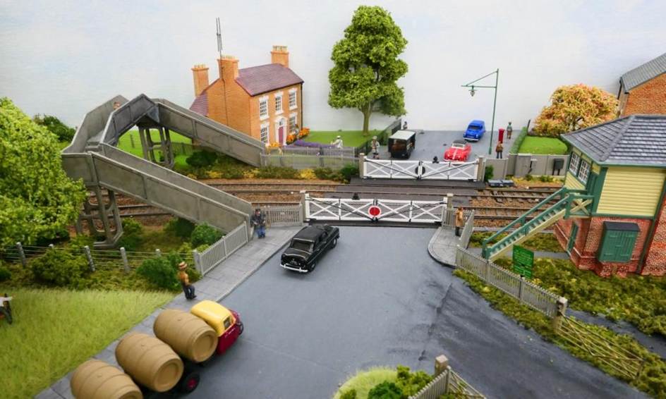 With its Mackenzie & Holland gates, Horsham Lane level crossing features a concrete footbridge which encroached into the front garden of Lavender House with its BBC H television ariel. To the left of the footbridge behind the bus stop a tree hides the scenic backboard with assists trains in disappearing from view. On the right part of the Signal Box is visible; this being now demoted to a Level Gate Crossing box. 
Below is virtually the same photograph shewing how a tree can significantly alter the view meaning the viewer now has to physically move to be able to see all.
www.EwhurstGreen.com 
