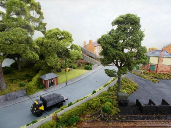 With its Mackenzie & Holland gates, Horsham Lane level crossing features a concrete footbridge which encroached into the front garden of  Lavender House  with its BBC  H  television ariel. To the left of the footbridge behind the bus stop a tree hides the scenic backboard with assists trains in disappearing from view. On the right part of the Signal Box is visible; this being now demoted to a Level Gate Crossing box. 
Below is virtually the same photograph shewing how a tree can significantly alter the view meaning the viewer now has to physically move to be able to see all.
www.EwhurstGreen.com  
