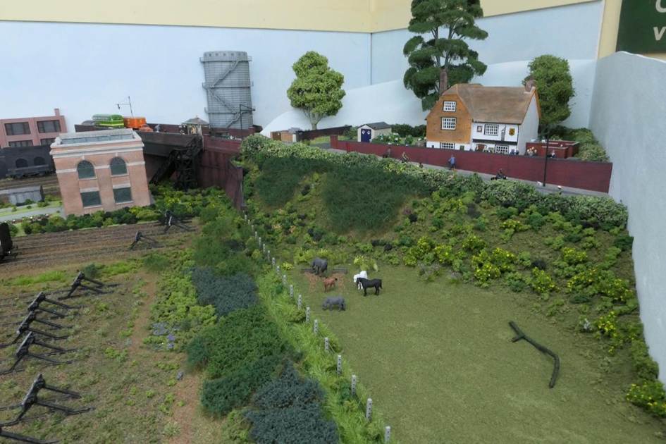 With a large tree still to be positioned in the field, the Woolpack Public House sits within an otherwise seemingly rural area on this side of the railway line. However, behind the Cathedral sub-station there is a modest agricultural factory on the far side of the railway bridge.
The thick dark patches of foliage are Lars op t Hof Scenery (in front of the fence and on the embankment side). Much of this scenery was supplied by Model Scenery Supplies of Cromer. 
www.EwhurstGreen.com 
